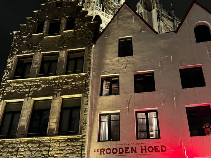 De gevel van Grand Café De Rooden Hoed bij avond, met daarachter de verlichte toren van de kathedraal