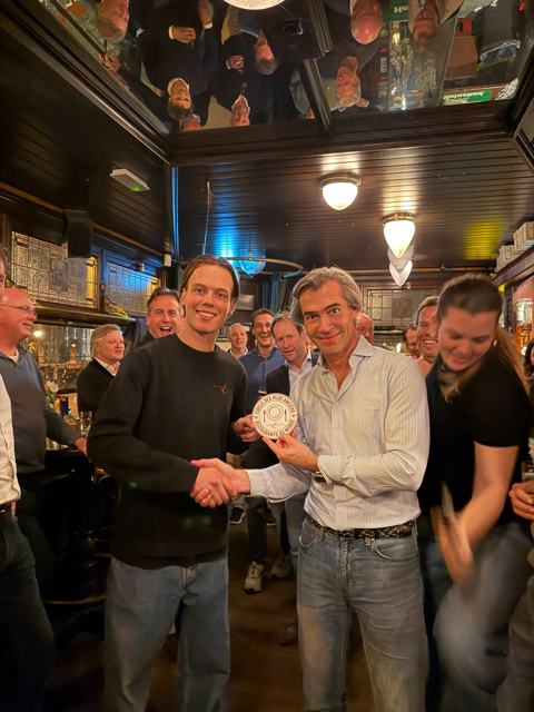 Twee mannen schudden elkaar de hand en houden samen het schild van de Cercle vast, omringd door lachende gasten