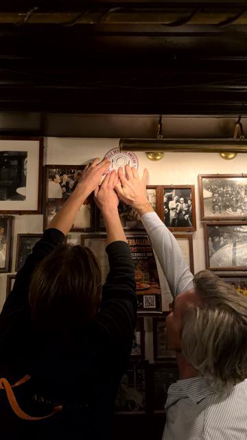 Drie handen drukken samen het ronde schild tegen de wand, tussen ingelijste zwart-witfoto's